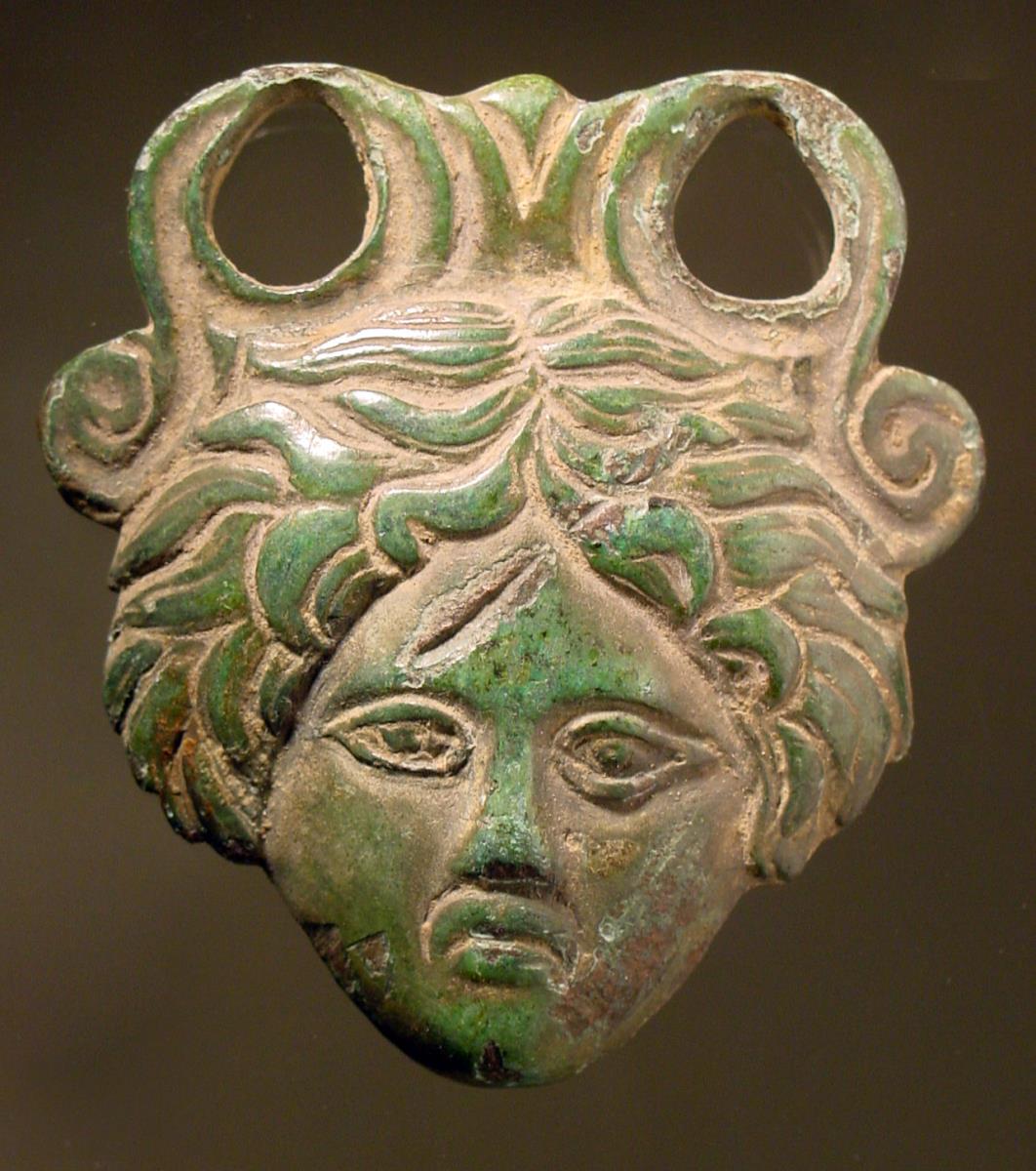 Henkelattasche eines Eimers "Beschlagstück in Form einer weiblichen Maske, wohl Gorgoneion, ... Oben Ansatz mit zwei großen Öffnungen und einer Volute jederseits. Hinten flach. Vollguß"
