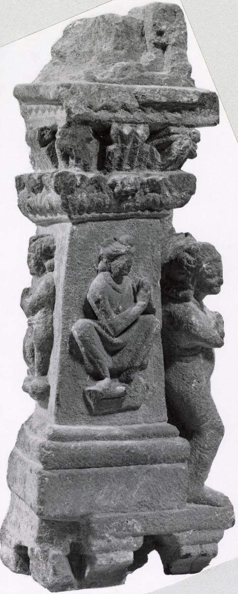 Eckpfeiler mit nach links gedreht sitzendem Bodhisattva in Verehrungsgeste (anjali-mudra) und anschließendem Fragmente einer Reliefszene zur einen Seite sowie nach rechts gedreht stehender Figur mit Turban zur anderen Seite
