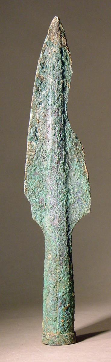 Speerspitze mit breiter flache Mittelrippe, auf der Längslinien zur Verzierung eingeritzt sind. Kleine Beschädigungen. In der Schafttülle Holzreste