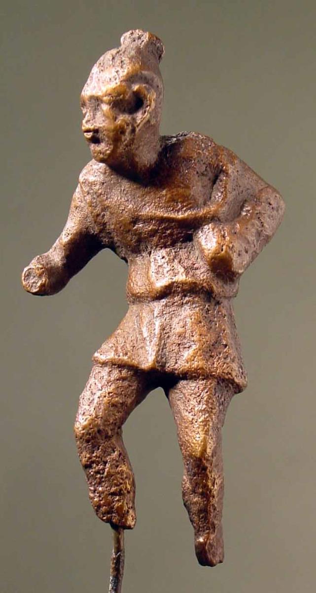 Römische Statuette eines Sklaven oder Possenreiβers (?). Kurzer Chiton, der von der linken Schulter niedergleitet. Rechte Hand mit sprechendem Gestus vorgestreckt. Kurzgeschnittene Haare mit Schopf. Beide Füße weggebrochen, r. Hand beschädigt. Rauhe Patina