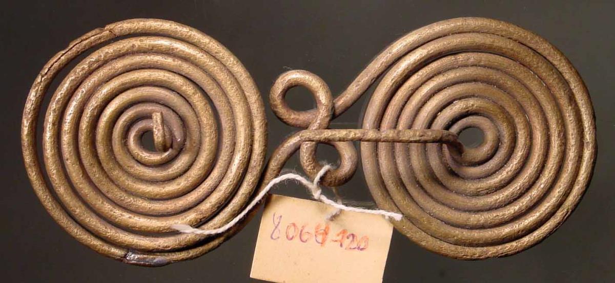 Brillenfibel. "Große Fibel aus Draht spiralförmig zusammengedreht. Das Ganze besteht aus einem und demselben Stück Draht, Nadel und Öse bilden Anfang und Ende der Windung. Stück der Nadel fehlt"