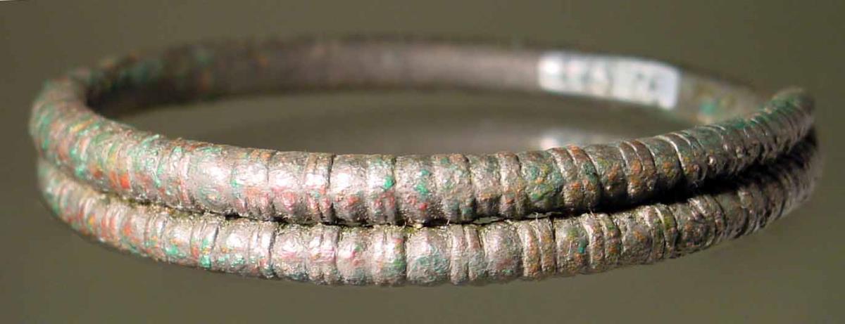 Armring aus starkem Draht mit übergreifenden Enden; in regelmäßigen Abständen folgen je zwei ringartige Anschwellungen aufeinander, sonst ohne Verzierung; der Draht verjüngt sich an den Enden