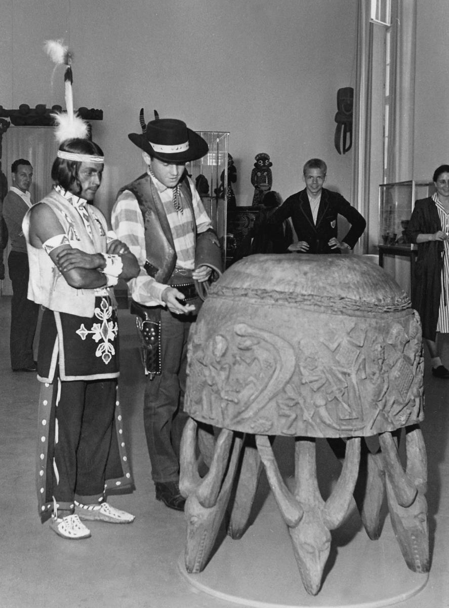"Der Indianer Sidi Borhara aus Utah und der Cowboy Koy Dunn aus Springfield bewundern eine grosse Trommel aus dem Grasland von Kamerun im M.f.v. Berlin. 13.07.1957"