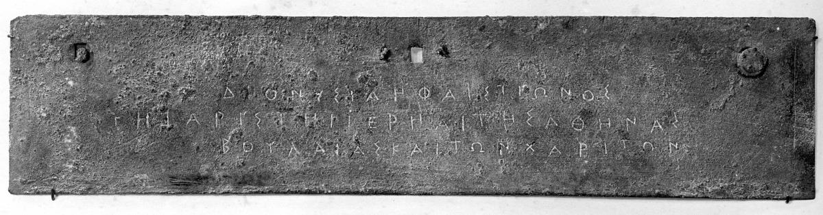 Dünne längliche Platte mit drei Nagellöchern und dreizeiliger griechischer Inschrift: ΔΙΟΝΥΣΙΑ ΗΦΑΙΣΤΙΩΝΟΣ / ΤΗΙ ΑΡΙΣΤΗΙ ΙΕΡΕΙΑΙ ΤΗΣ ΑΘΗΝΑΣ / ΒΟΥΛΑΙΑΣ ΚΑΙ ΤΩΝ ΧΑΡΙΤΩΝ