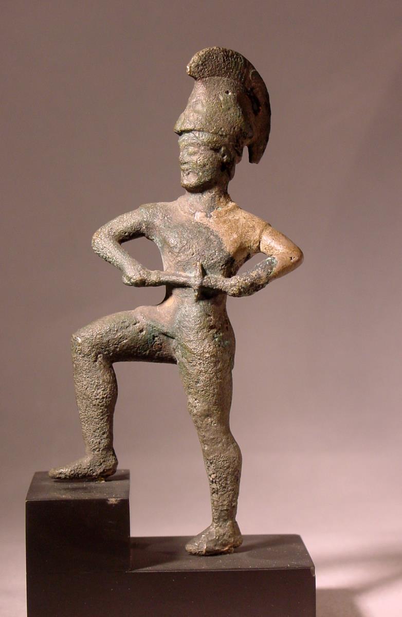Archaische Statuette eines Kriegers mit Panzer, Beinschienen und hohem Helm, das rechte Bein hoch gehoben, das Schwert ziehend