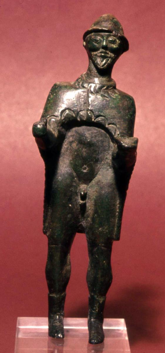 Archaische Votivstatuette eines stehenden, mit Mantel, Hut und Schuhen bekleideten Mannes