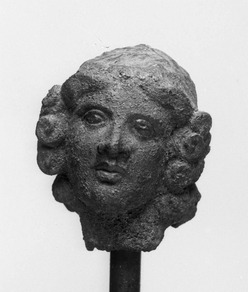 Köpfchen eines Knaben, wohl Eros, von einer Statuette abgebrochen, Haarenden in Schneckenlocken ausgehend, breite Haarbinde, Hohlguß