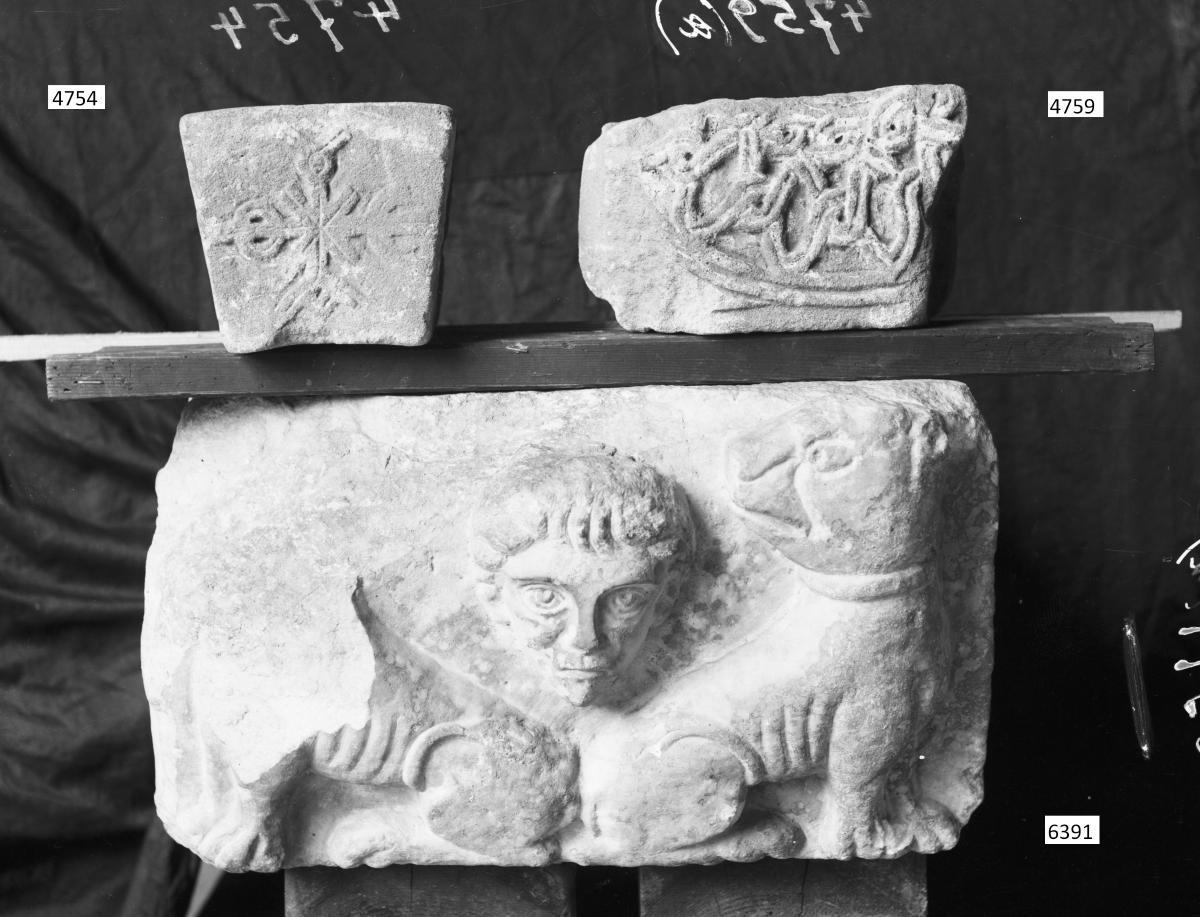 Kubisches Architekturelement mit Relief eines männlichen Kopfes zwischen zwei zurückblickenden Löwen